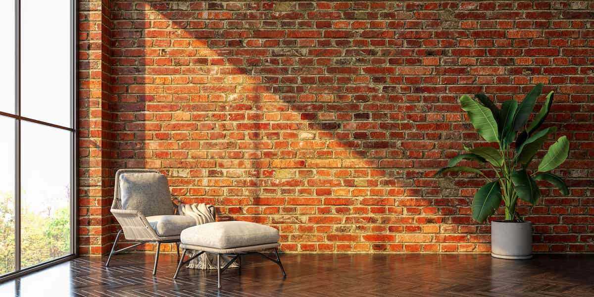 A modern living room with exposed brick walls, sleek apartment flooring in a herringbone pattern, a cozy chair with an ottoman, and a potted plant by a large window.