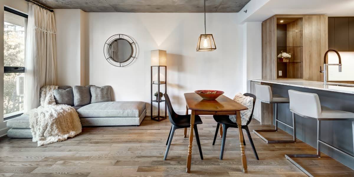 Open-plan apartment living and kitchen with warm wood plank flooring for apartments, gray sectional sofa, dining table, and breakfast bar stools.